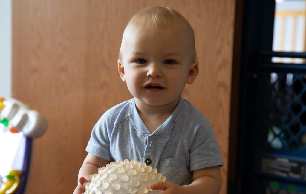 A toddler standing staring at the camera holding a ball