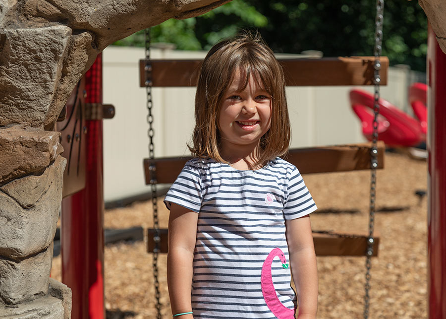 A child smiling at the camera standing on the red and brown playground