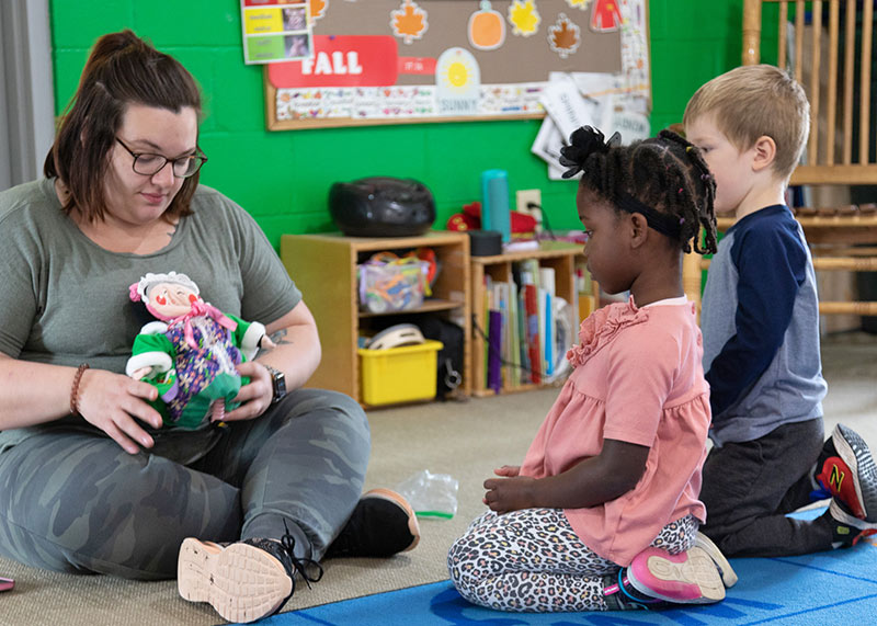 A teacher sitting on the floor holding a baby doll with two students watching